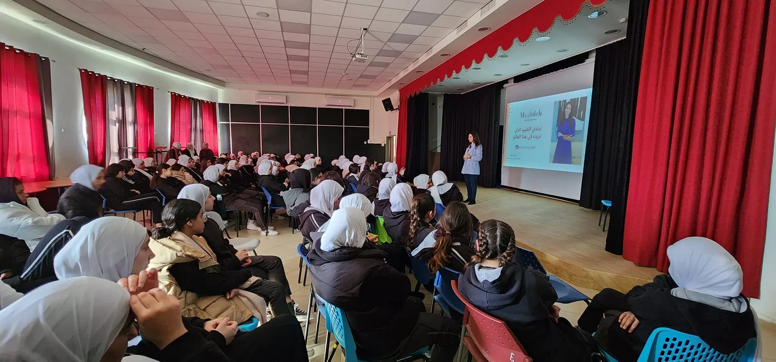 תלמידות יושבות באולם עם במה ומסך, והמרצה לצד המסך