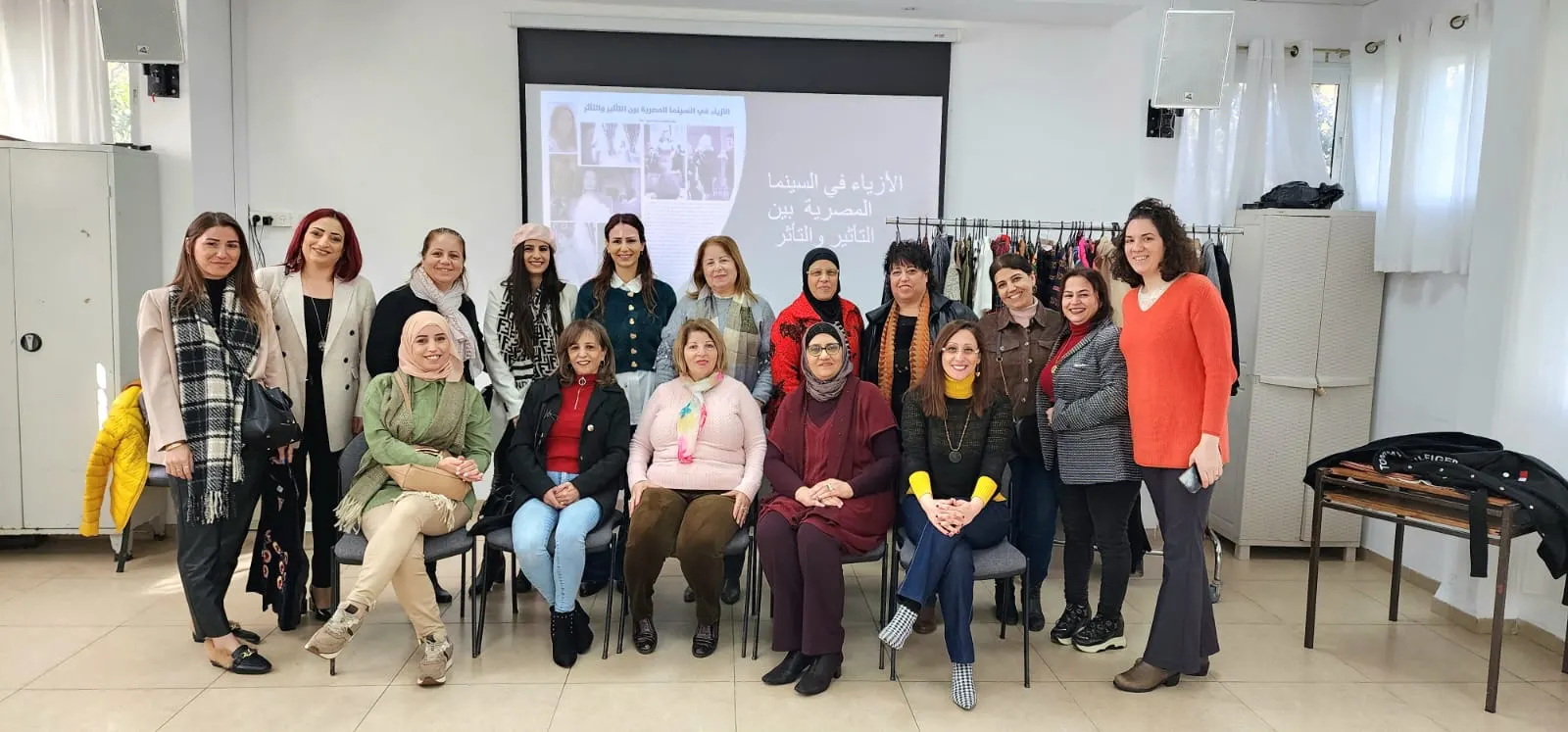 קבוצת נשים יושבת ועומדת באולם, מאחוריהן מתלה בגדים ומסך מצגת